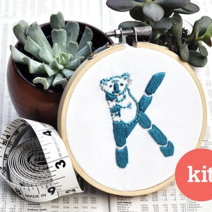 May include: A white embroidery hoop with a teal blue embroidered letter K with a koala bear inside the letter. The hoop is on a white surface with a measuring tape and a succulent plant in a brown pot.
