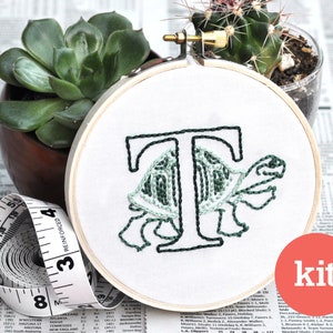 May include: A green and black embroidery kit featuring a turtle with the letter "T" on its shell. The embroidery is in a wooden hoop and the word "kit" is printed on a red circle.