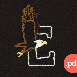 Könnte beinhalten: Gestickte Darstellung eines Weißkopfseeadlers im Flug, dessen Flügel die Form des Buchstabens "E" in weißem Garn auf einem schwarzen Stoffhintergrund bilden.