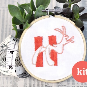 May include: A DIY embroidery kit featuring a pink bunny rabbit with a red and white striped background. The kit includes a hoop, fabric, and embroidery floss. The word "kit" is printed in red on a white circle.