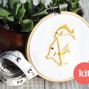 May include: A DIY embroidery kit featuring a white embroidery hoop with a yellow embroidered letter "F" with two fish designs. The kit includes a white fabric, embroidery floss, and a needle. The word "kit" is printed in red on a white circle.