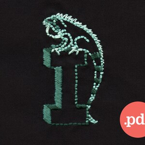 May include: Green embroidered design of a lizard on a black fabric background. The lizard is curled around the letter "I" which is also embroidered in green.
