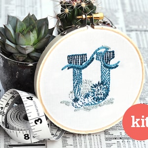 May include: An embroidery hoop with the letter "U" stitched in shades of blue. The word "kit" is in a red circle. A measuring tape and potted succulents are in the background.