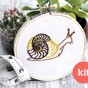 May include: A DIY embroidery kit featuring a snail with a brown and yellow shell. The kit includes a hoop, fabric, and embroidery floss. The word "kit" is printed in white on a red circle.