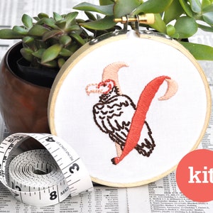 May include: Embroidery kit featuring a red and brown embroidered letter V with a stylised vulture design. The embroidery is on a white fabric stretched over a wooden embroidery hoop. The hoop is 15 cm in diameter. The word "kit" is printed on a red circle.