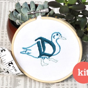 May include: An embroidery kit featuring a stylised bird with the letter "D" incorporated into its body, embroidered in blue and teal. The embroidery is framed in a wooden hoop and the word "kit" is visible in the lower right corner.