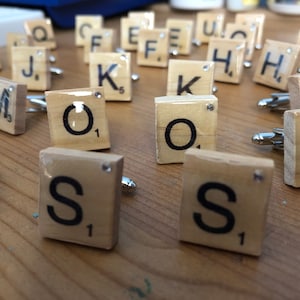 May include: A set of wooden cufflinks featuring letters of the alphabet, each letter is set in a square tile with a black border and a white background. The tiles are made to look like Scrabble tiles.