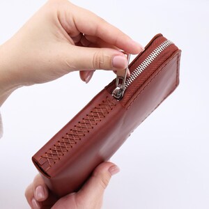 May include: A brown leather zipped pouch with a decorative stitch pattern. The zipper is open and the pouch is being held by a hand.