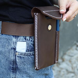 A brown leather phone case with a white stitch detail. The case has a flap that closes with a snap button. The case is attached to a brown leather belt.Genuine Leather Phone Custom Case With Belt Clip, Leather Pen And Phone Holster, Individually Phone Size Leather Case With Metal Belt Clip