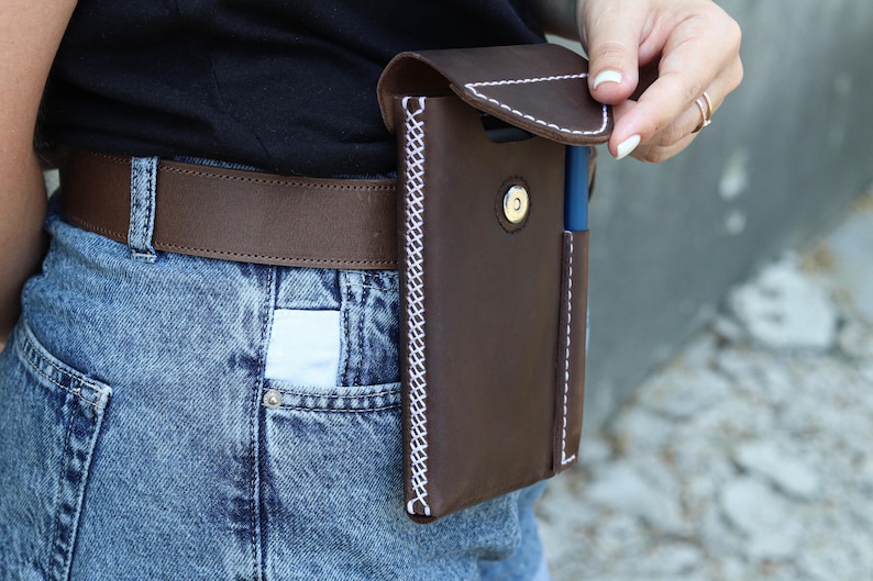 A brown leather phone case with a white stitch detail. The case has a flap that closes with a snap button. The case is attached to a brown leather belt.Genuine Leather Phone Custom Case With Belt Clip, Leather Pen And Phone Holster, Individually Phone Size Leather Case With Metal Belt Clip