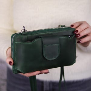 以下が含まれることがあります： 滑らかなマット仕上げのダークグリーンの長方形クロスボディバッグ。上部にジッパー開閉式、前面にはフラップとスナップボタンで固定されたポケットが付いています。シルバーカラーの金属製金具と、肩掛け用の細いストラップが特徴です。