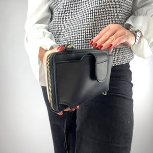 以下が含まれることがあります： 長方形の黒いレザー製クロスボディバッグ。側面にはゴールドトーンの金属製ジッパー開閉部があり、前面にはタブ留め付きのフラップポケットが付いています。バッグには細い黒のストラップと、縁に沿って見えるステッチが施されています。