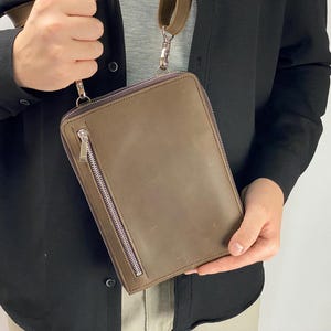 以下が含まれることがあります： オリーブブラウンのレザー製長方形クロスボディバッグ。前面に縦型のシルバーカラーのファスナーポケット、上部にメインのファスナー開閉部を備え、シルバーカラーの金属製クリップで取り付けられた同色の調節可能なショルダーストラップが付いています。