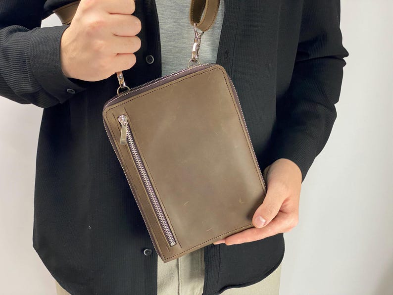 以下が含まれることがあります： オリーブブラウンのレザー製長方形クロスボディバッグ。前面に縦型のシルバーカラーのファスナーポケット、上部にメインのファスナー開閉部を備え、シルバーカラーの金属製クリップで取り付けられた同色の調節可能なショルダーストラップが付いています。