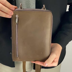 以下が含まれることがあります： トープ色のレザー製長方形クロスボディバッグ。前面に縦型のファスナーポケット、上部にファスナー開閉口、シルバーカラーの金属製金具を備えています。バッグの上部にはストラップを取り付けるための2つのアタッチメントポイントがあります。