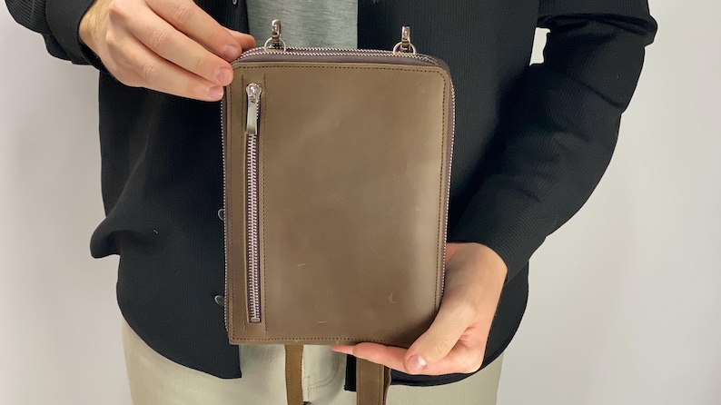 以下が含まれることがあります： トープ色のレザー製長方形クロスボディバッグ。前面に縦型のファスナーポケット、上部にファスナー開閉口、シルバーカラーの金属製金具を備えています。バッグの上部にはストラップを取り付けるための2つのアタッチメントポイントがあります。