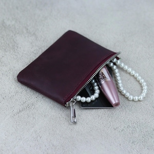 May include: A burgundy leather zipped pouch with a silver zipper pull. The pouch is open and contains a black card, a pearl necklace, and a pink mascara tube with the text "L'Oreal Paris Voluminous Lash Paradise Mascara" printed on the side.