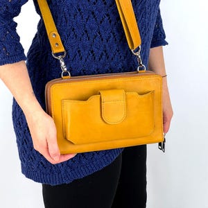 May include: Mustard yellow leather crossbody bag with a front pocket and a flap closure. The bag has a long, adjustable strap and a zipper closure. The bag is held by a person wearing a blue sweater.