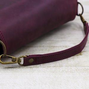 May include: A close-up of a burgundy leather handbag with a strap and a brass clasp. The bag is on a light wood surface.