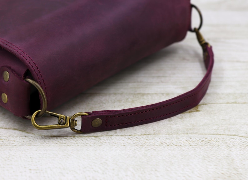 May include: A close-up of a burgundy leather handbag with a strap and a brass clasp. The bag is on a light wood surface.