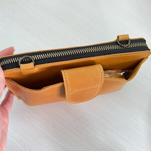 以下が含まれることがあります： マスタードイエローのレザー製クロスボディバッグ。上部にダークカラーの金具付きファスナー、ストラップ取り付け用の金属製Dリング2つ、スナップボタン付きフラップで固定されたフロントポケットを備え、マットな質感で仕上げられています。