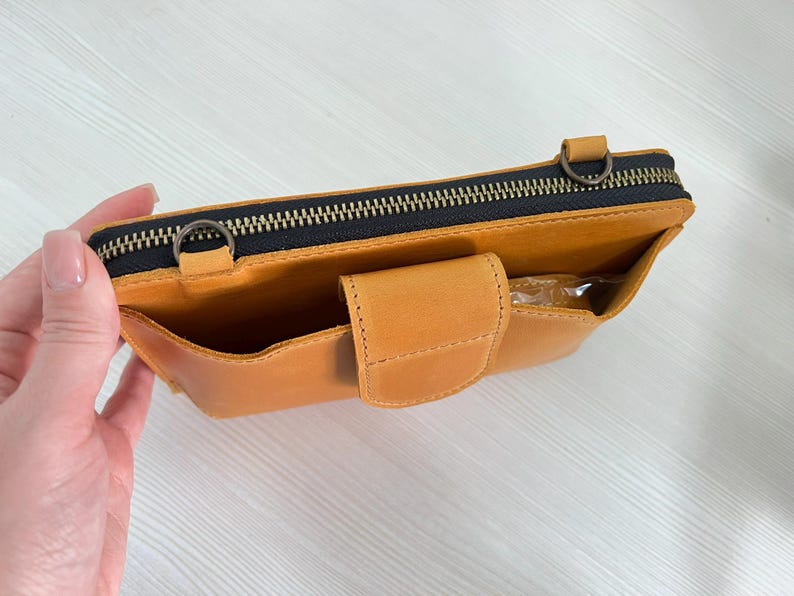 以下が含まれることがあります： マスタードイエローのレザー製クロスボディバッグ。上部にダークカラーの金具付きファスナー、ストラップ取り付け用の金属製Dリング2つ、スナップボタン付きフラップで固定されたフロントポケットを備え、マットな質感で仕上げられています。