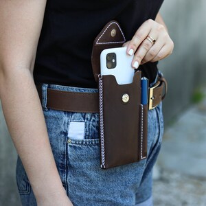 A brown leather phone case with a white stitching detail. The case is attached to a brown leather belt with a gold buckle. A blue pen is in a separate pocket on the case. Genuine Leather Phone Custom Case With Belt Clip, Leather Pen And Phone Holster, Individually Phone Size Leather Case With Metal Belt Clip