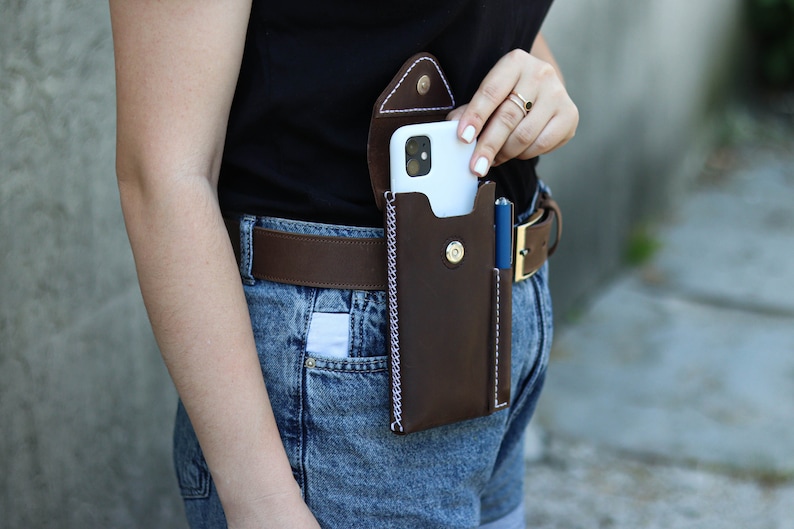 A brown leather phone case with a white stitching detail. The case is attached to a brown leather belt with a gold buckle. A blue pen is in a separate pocket on the case. Genuine Leather Phone Custom Case With Belt Clip, Leather Pen And Phone Holster, Individually Phone Size Leather Case With Metal Belt Clip
