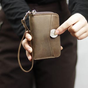 May include: A brown leather wallet with a zip fastening and a wrist strap. The wallet has a coin pocket with a silver coin visible.