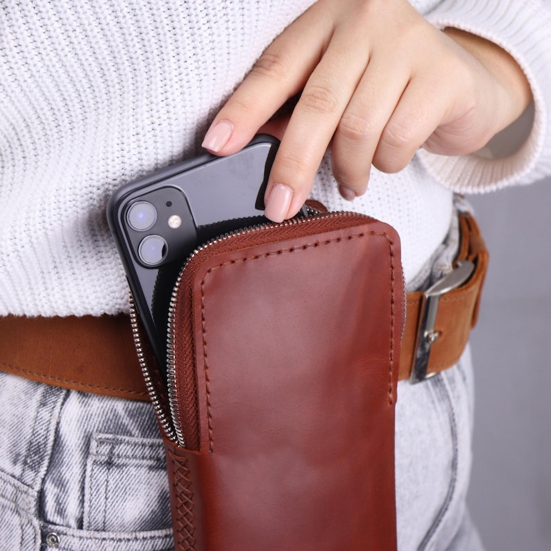 May include: A brown leather phone case with a zip closure. The case is being worn on a belt and a smartphone is being inserted into the case.