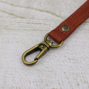 May include: A close-up of a brown leather strap with a brass swivel clasp. The strap has a stitched design and a small brass button.
