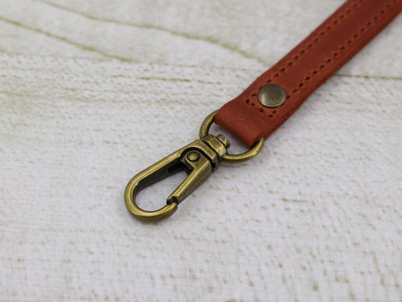 May include: A close-up of a brown leather strap with a brass swivel clasp. The strap has a stitched design and a small brass button.