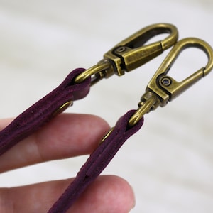 May include: Two burgundy leather straps with gold metal clasps. The straps are attached to the clasps with stitching.