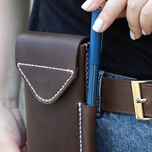 A brown leather phone case with white stitching. The case has a flap that closes with a snap. A blue pen is inserted into the case.