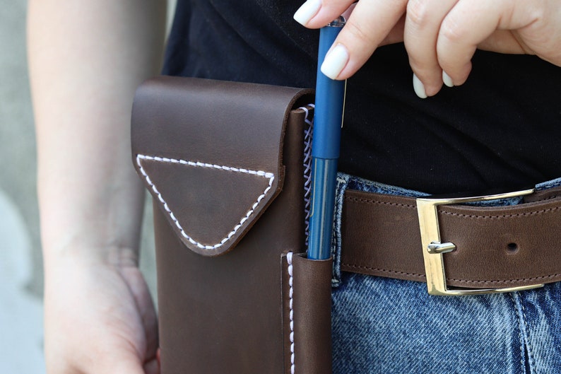 A brown leather phone case with white stitching. The case has a flap that closes with a snap. A blue pen is inserted into the case.