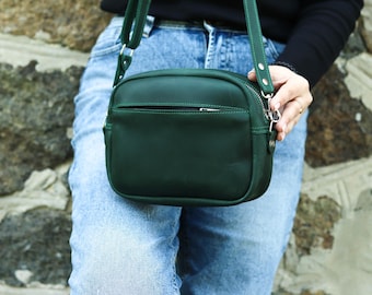 Puede incluir: Un bolso bandolera de cuero verde oscuro con cremallera superior y correa ajustable. El bolso es rectangular con esquinas redondeadas y un pequeño bolsillo frontal. La correa y el bolso son del mismo color. El bolso lo lleva una persona con vaqueros azules.