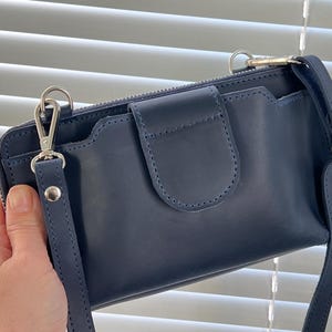 以下が含まれることがあります： 滑らかな質感のネイビーブルーのクロスボディバッグ。中央にはステッチが施されたフラップ式の留め具があり、上部にはファスナーが付いています。バッグの両サイドには、シルバーカラーの金属製クリップで取り外し可能なショルダーストラップが取り付けられています。