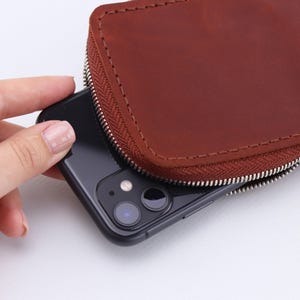 May include: A brown leather phone case with a zipper closure. A black smartphone is partially visible inside the case.