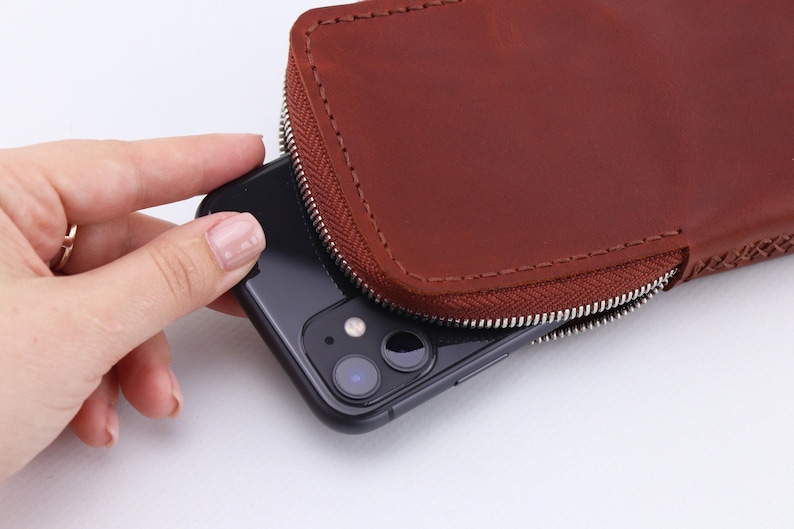 May include: A brown leather phone case with a zipper closure. A black smartphone is partially visible inside the case.