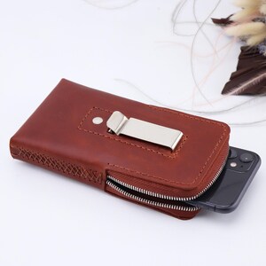 May include: A brown leather phone case with a zipper closure and a metal clip. The case has a stitched design and is holding a black smartphone.