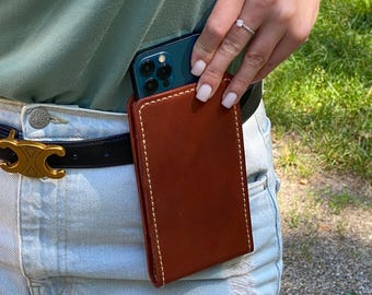 May include: A rectangular brown leather phone holster with visible white stitching along the edges. The holster is attached to a black belt with a gold-toned buckle, worn over light blue denim jeans and a sage green top.