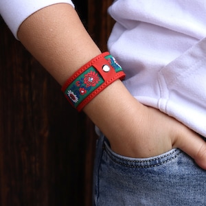 Könnte beinhalten: Rotes Lederarmband mit einem grün und floral bestickten Band. Das Armband wird mit einem silbernen Druckknopfverschluss befestigt. Das Armband ist etwa 3,8 cm breit.