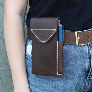 A brown leather phone case with a white stitch design. The case is attached to a brown leather belt with a gold buckle. The case has a separate pocket for a pen. Genuine Leather Phone Custom Case With Belt Clip, Leather Pen And Phone Holster, Individually Phone Size Leather Case With Metal Belt Clip