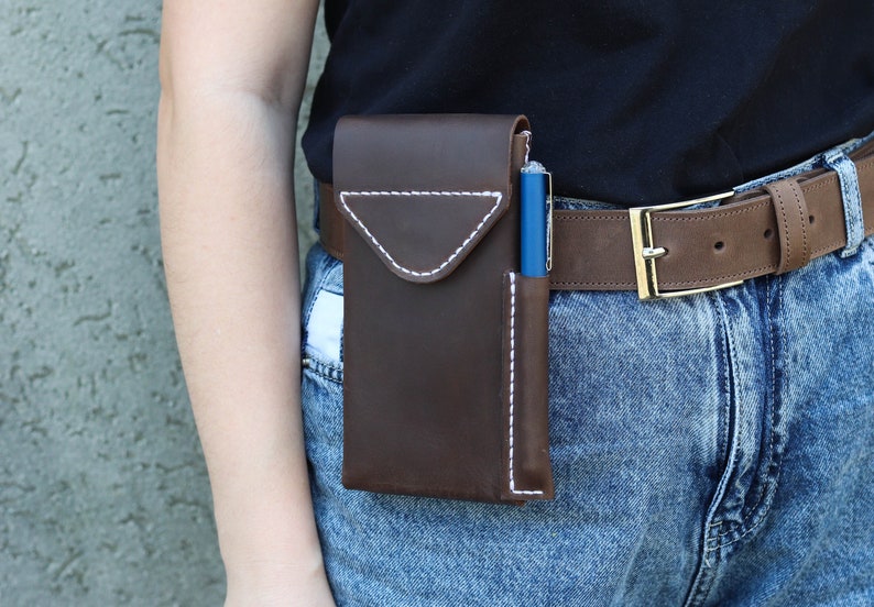 A brown leather phone case with a white stitch design. The case is attached to a brown leather belt with a gold buckle. The case has a separate pocket for a pen. Genuine Leather Phone Custom Case With Belt Clip, Leather Pen And Phone Holster, Individually Phone Size Leather Case With Metal Belt Clip