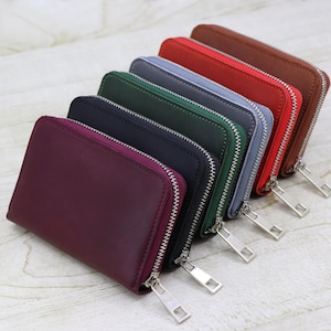 Puede incluir: Una colección de carteras con cremallera de colores en fila. Las carteras son de color burdeos, azul marino, verde oscuro, gris, rojo y marrón. Cada cartera tiene una cremallera y un tirador plateados.