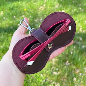 Puede incluir: Un estuche para gafas de cuero burdeos con cierre de presión y un gancho de metal. El estuche contiene unas gafas de sol rosas.