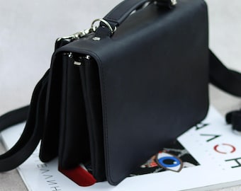 May include: A black leather satchel bag with a top handle and a long adjustable strap. The bag has a flap closure and multiple compartments.