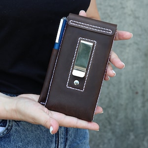 A brown leather phone case with a silver metal clip and a pen holder. The case has white stitching around the edges.
