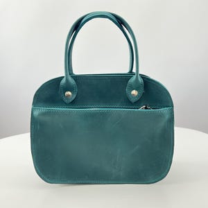 May include: Teal handbag with a rounded shape and two top handles. The bag has a front pocket with a zip and is made of a smooth material. The handles are attached with gold-coloured rivets. The bag is on a white surface.
