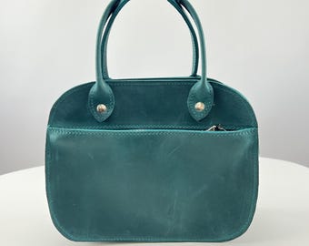 May include: A teal leather handbag with a rounded top, two fixed handles, and a horizontal zippered pocket on the front. The bag features silver-toned metal rivets securing the handles to the main body, providing a structured appearance.